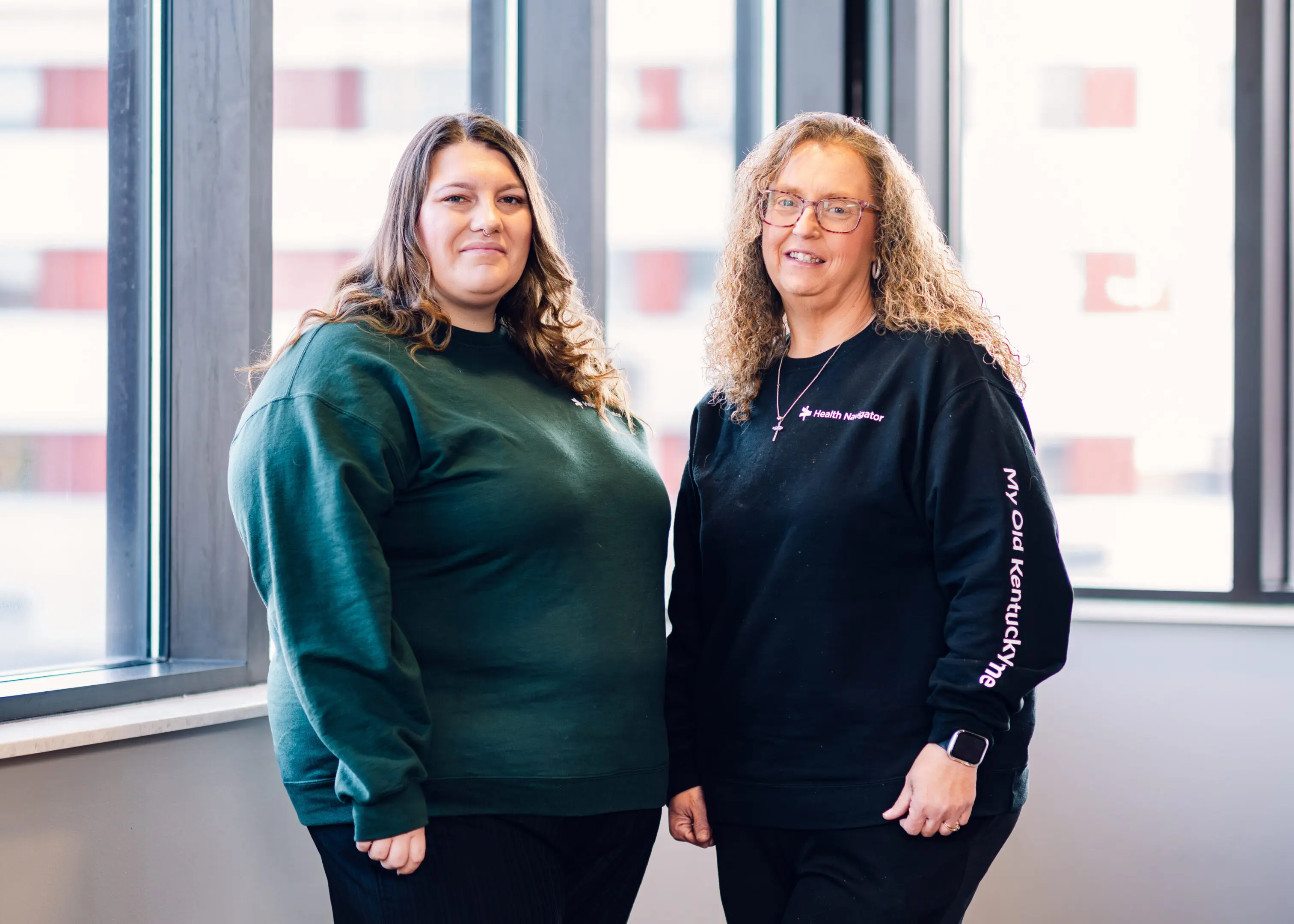 Two women standing indoors, smiling and dressed in casual sweatshirts.