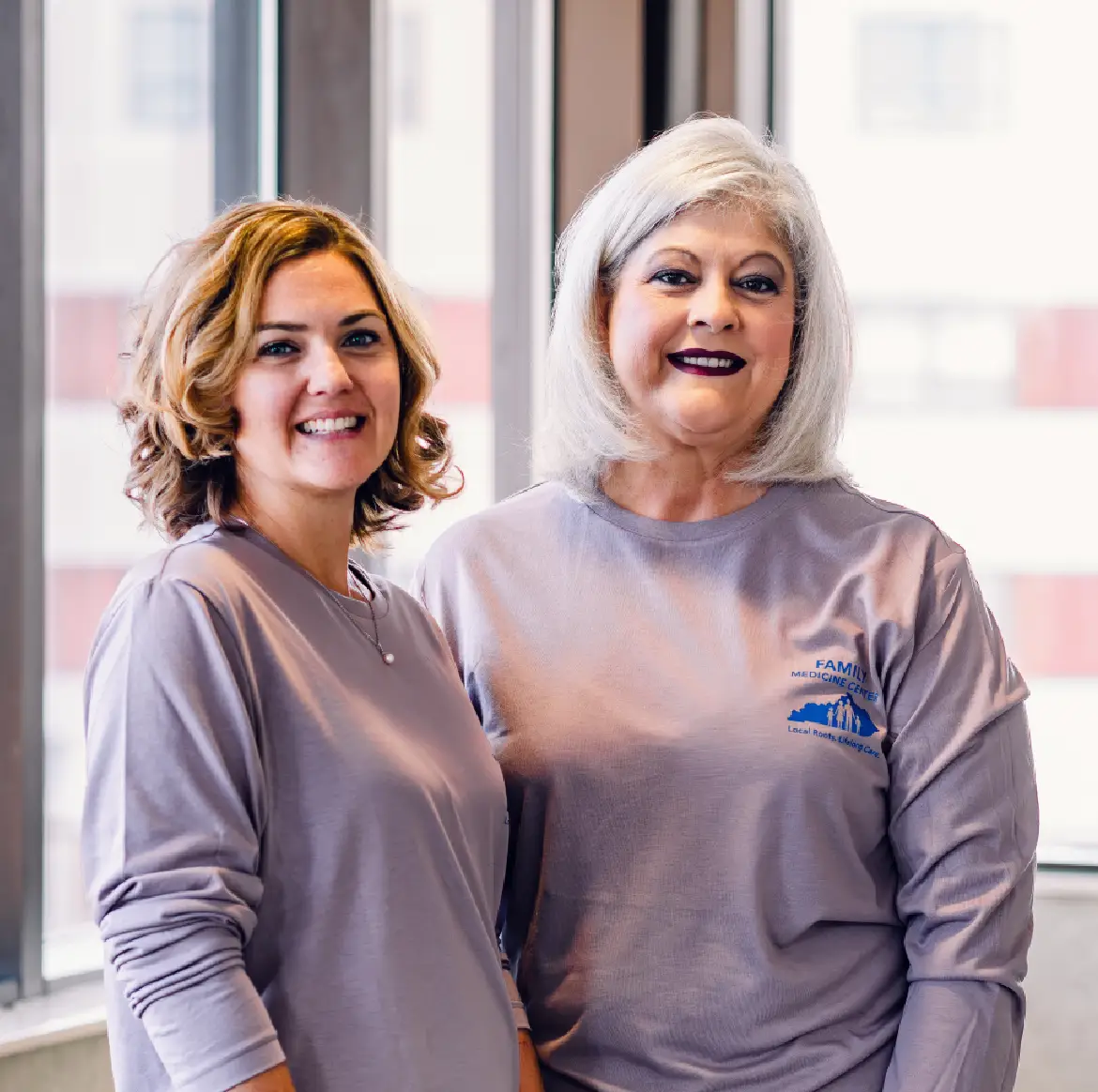 Two smiling women wearing matching lavender tops indoors.